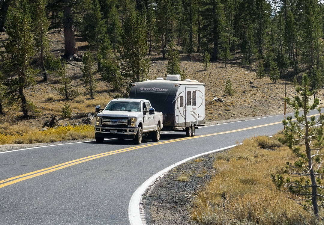 Toy Hauler A white pickup truck towing a travel trailer with "EverGreen" written on it travels along a two-lane road with a double yellow line. The road is surrounded by trees and dry grass, suggesting a rural or forested area.