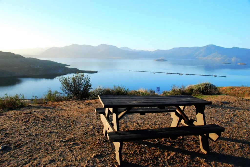 Scenic view of a mountain-surrounded lake, with a picnic table at the edge of the land and water.