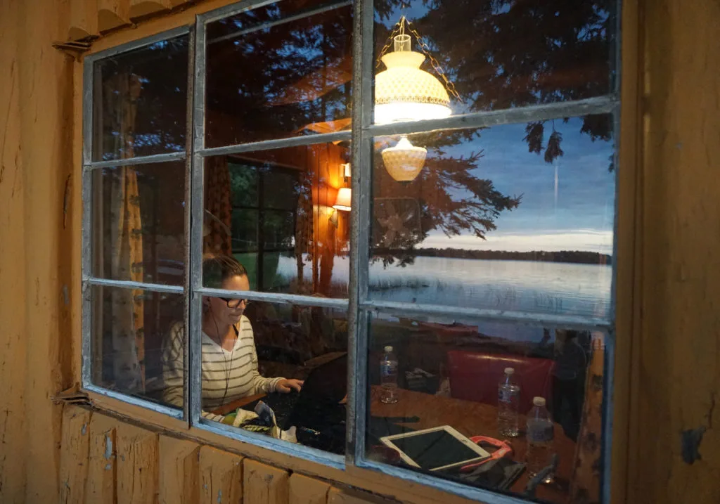 View through a rustic cabin window of a woman working on a laptop at a kitchen table.
