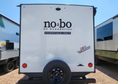 The rear view of a white No Boundaries (NOBO) travel trailer. The trailer features the "NOBO" logo, the phrase "no boundaries ESSENTIALS ONLY," and a social media handle and icons for Instagram and YouTube toward the lower right side of the trailer.