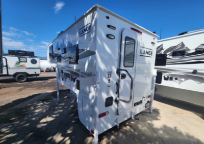 A white Lance camper is parked on a concrete surface on a sunny day. The camper features multiple windows and storage compartments, with the word "LANCE" prominently displayed on the back and side. Other campers and vehicles are visible in the background.