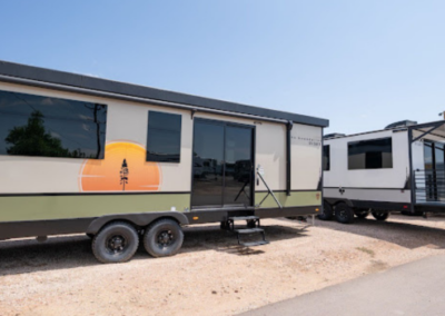 Two modern travel trailers are parked on a gravel lot under a clear blue sky. One trailer features a stylized tree emblem on its side and a large glass door with steps leading up. The second trailer, mostly white with black accents, is positioned behind the first.