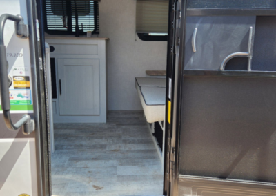 This image shows the interior view of an RV through its open door. Inside, there is light wood plank flooring, a white cabinet, and seating with a cushion on the right side. Sunlight filters in from a window with a blind. The door has a screen attached.