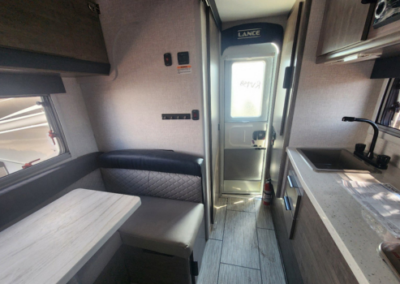 Interior of a camper showing a small dinette with cushioned seating and a table on the left, and a compact kitchen on the right with a countertop, sink, and faucet. A door with a window is at the far end, and sunlight filters through, illuminating the space.