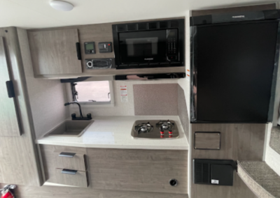 The image shows a compact RV kitchen with a sink, a two-burner propane stove, and a microwave above. Next to the microwave is a small refrigerator. The kitchen has a modern design with light wood cabinetry and a clean, organized layout.