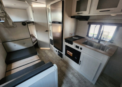 Interior of a compact camper van showing a small kitchen area with a sink, stovetop, and cabinets. Adjacent to the kitchen is a small bunk bed area with a curtain. The seating area with cushioned benches is opposite the kitchen and is furnished with a foldable table.