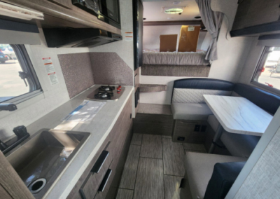 A compact RV interior featuring a kitchenette with a sink and stovetop on the left, a small dining area with a marble-patterned table and cushioned seats on the right, and a bed at the back. Neutral tones of gray and beige dominate the space.