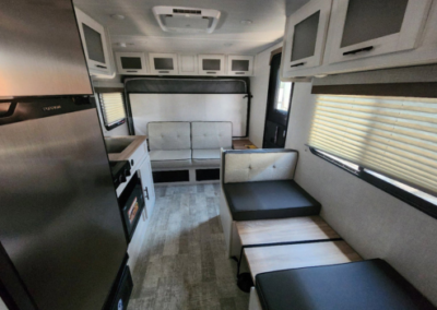 The interior of a small, modern RV featuring a compact kitchen with a fridge and oven on the left, opposite a bench seating area. The rear of the RV has additional seating and storage. Light-colored cabinetry and large windows create a bright ambiance.