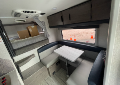 Interior of a modern RV featuring a table with cushioned seats on either side, a window above the seating area, and overhead storage cabinets. The back part of the RV has a sleeping area with a box on the bed. Orange traffic cones are visible outside the window.