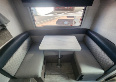 A compact RV dining area with a small rectangular table surrounded by U-shaped cushioned seating. The cushions have a quilted pattern on the backrests and plain seats. A window behind the seating provides natural light and an outside view.