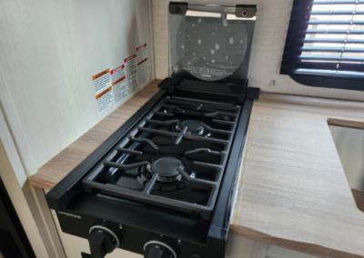 Image of a compact kitchen stove with two gas burners and a glass cover, set into a countertop. Control knobs are located at the front of the stove, and there are warning labels on the wall next to it. Sunlight is coming in through a window with blinds.