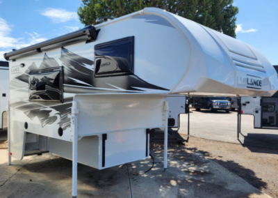 A Lance truck camper is parked outdoors on concrete. The camper has a white exterior with black and gray mountain graphics on the side, a raised over-cab sleeping area, and a retractable awning. Trees and other RVs are visible in the background under a partly cloudy sky.