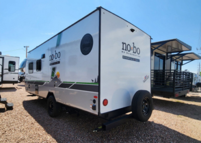 A white No Boundaries travel trailer is parked on gravel next to other trailers. The trailer has a graphic of mountains and a sunset on the side, with a spare tire mounted on the rear. A modern building with a porch is visible in the background.