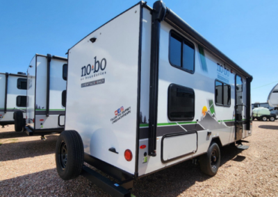 A compact, white travel trailer with the brand "no•bo by Forest River" displayed on the back, parked in an outdoor lot among other trailers. The trailer has graphic designs of mountains, trees, and a sunset on its side, and the hashtag #NOBOGOAT on its back.