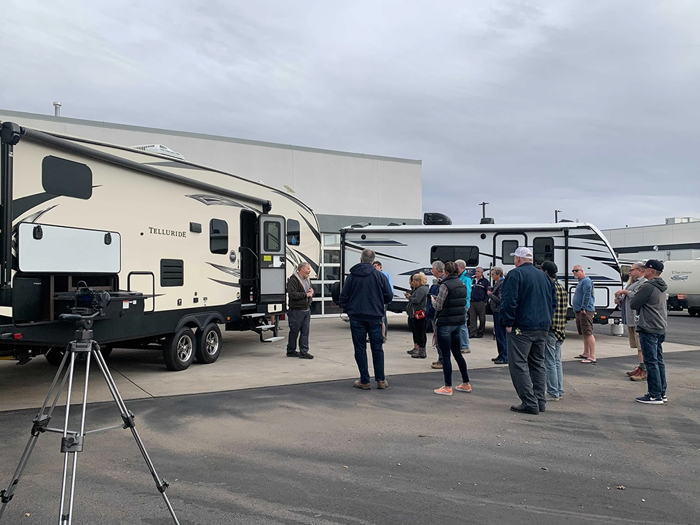 A group of people stand outdoors in front of two parked RVs, listening to a man giving a presentation. One person operates a camera on a tripod, while others watch and listen attentively. The sky is overcast, and a large building is in the background.