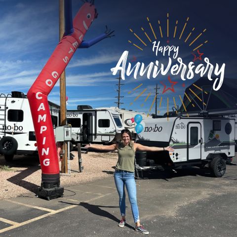 A woman stands smiling with arms outstretched in front of RV trailers, next to a red inflatable tube man labeled "CAMPING." "Happy Anniversary" is written above her with a burst of yellow and red lines.