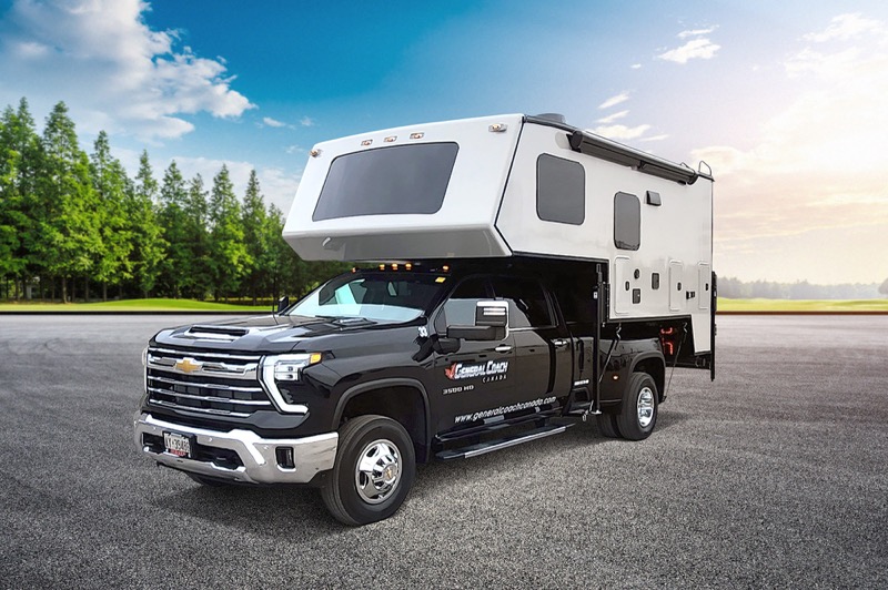 A black Chevrolet pickup truck with a large white camper attached is parked on a paved surface. There are green trees and a blue sky with clouds in the background.