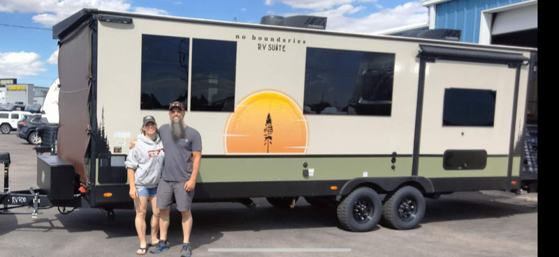 A smiling couple stands in front of a large beige and green "No Boundaries RV Suite" travel trailer with black trim, parked on a lot under a partly cloudy sky.