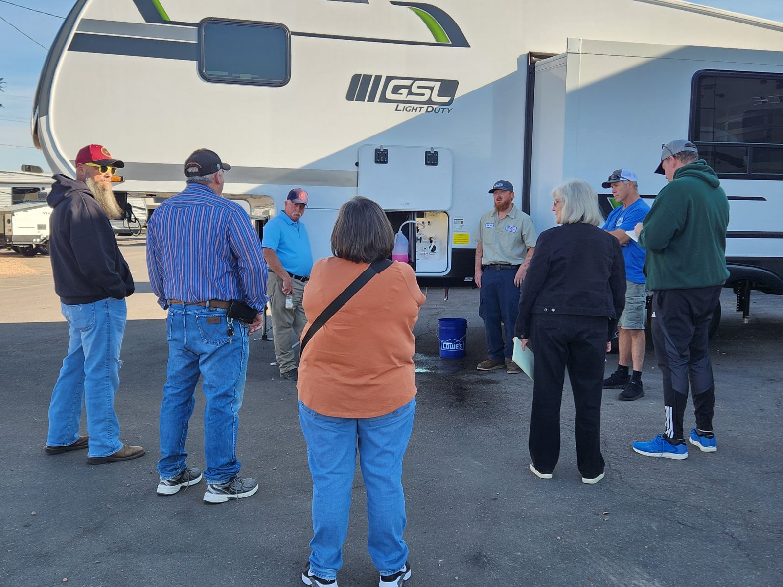 A group of nine adults stands in a circle outside near a large white RV, engaged in conversation on a sunny day. Most are wearing casual clothes and hats. One man appears to be explaining something to the group.