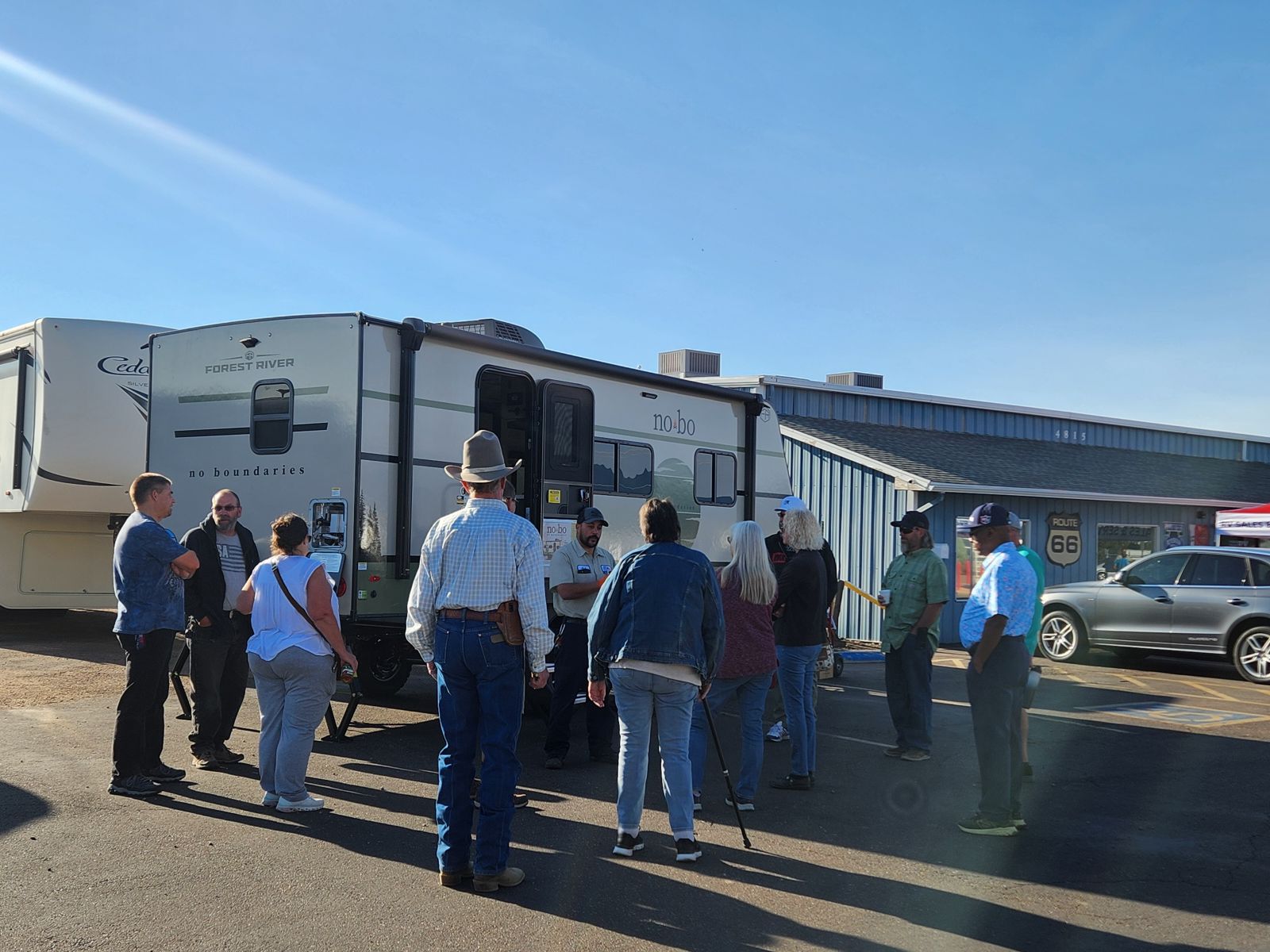 A group of people stands outside near an RV and a building under a clear blue sky, listening to a person speaking. Some people are wearing hats and casual clothes, and cars are parked nearby.