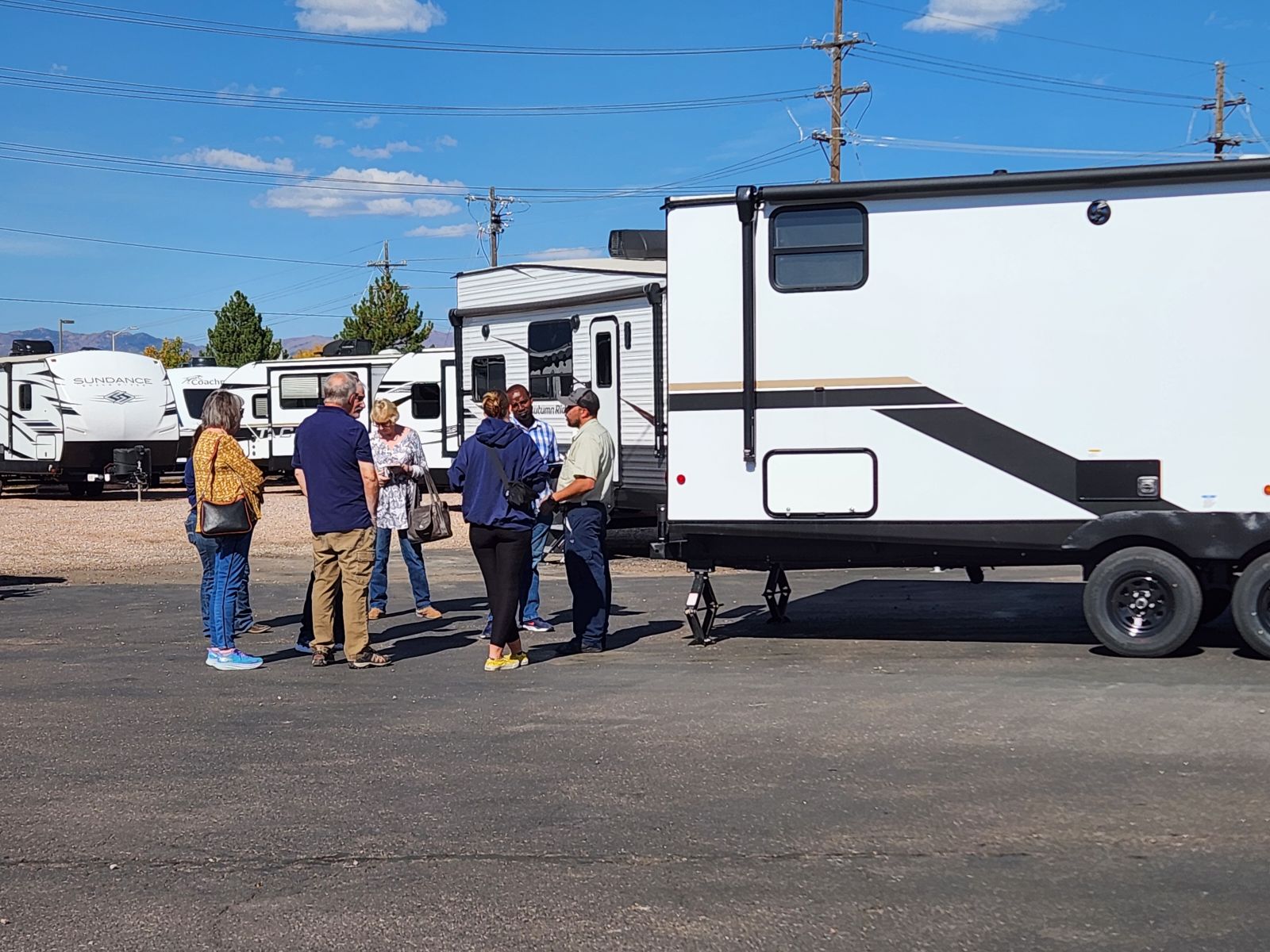 A group of six people stand in a parking lot next to a white travel trailer, engaged in conversation. Several other RVs and campers are visible in the background under a clear blue sky.