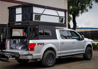 A silver pickup truck with an extended pop-up camper in the truck bed is parked beside a building, with the camper’s side windows visible.