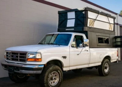 A white pickup truck with an elevated pop-up camper shell is parked next to a beige building with a maroon stripe. The camper’s side window is open, and the truck appears ready for camping or travel.