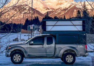 A gray pickup truck with a pop-up camper parked on a snowy road, with snow-covered mountains and pine trees in the background.