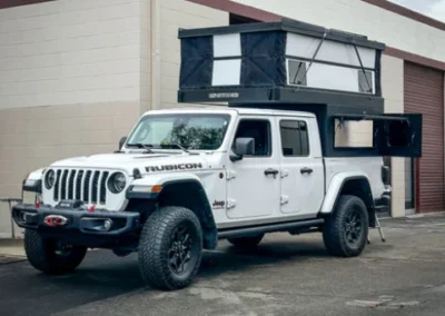 A white Jeep Rubicon with a rooftop tent and pop-up camper attachment is parked outside a building with a garage door.