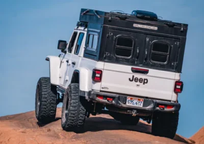 A white Jeep with large off-road tires climbs over rugged rocks under a clear blue sky, equipped with a black camper on the back.