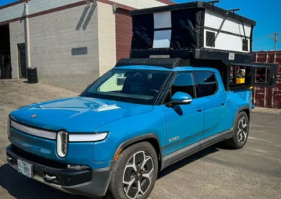 A blue Rivian electric pickup truck with a black and white pop-up camper attached to its bed is parked outside a building on a sunny day.