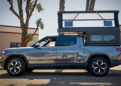 A gray Rivian pickup truck with a black and white camper attachment on the bed is parked on a street next to a tree and building, with the camper roof popped up.