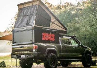 A black pickup truck with an extended rooftop tent is parked outdoors. The tent is open and elevated, and red fuel containers are mounted on the side. Trees and greenery surround the vehicle.