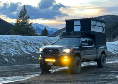 A dark pickup truck with a rooftop tent is parked on a muddy road, surrounded by snow-covered hills and pine trees at sunset, with mountains in the background.