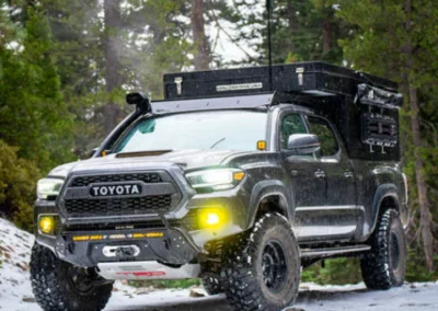 A lifted Toyota Tacoma with off-road tires and camping gear drives on a snowy forest trail, surrounded by tall pine trees.