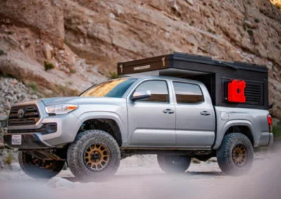 A silver pickup truck with large off-road tires and a black rooftop camper is parked on rocky terrain near a canyon wall. A red fuel canister is mounted on the camper.