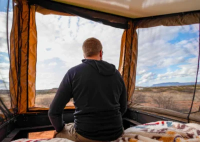 A person sits inside a tent, facing away and looking out at a scenic desert landscape under a partly cloudy sky. Blankets with colorful patterns cover the surface inside the tent.