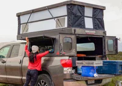 A person in a red shirt and hat opens the side hatch of a gray pickup truck with a pop-up camper in the bed. Camping supplies are visible, and the scene is outdoors.