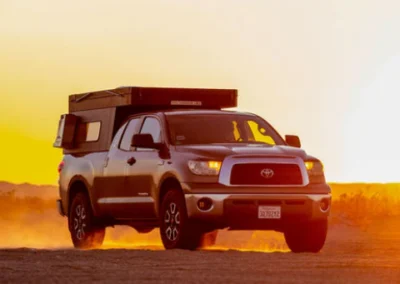 A silver Toyota pickup truck with a camper shell drives on a dusty road at sunset, with an orange sky and distant hills in the background.