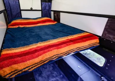 A cozy bed with a blue blanket and red-orange stripes inside a compact camper or van with white and black walls.
